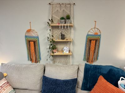 A macramé shelf with small plants and a clock above a gray couch with blue and orange pillows